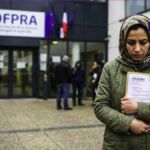 Une femme tenant un dossier de demande d'asile avec un tampon "Refus" devant les bureaux de l'Ofpra en France.